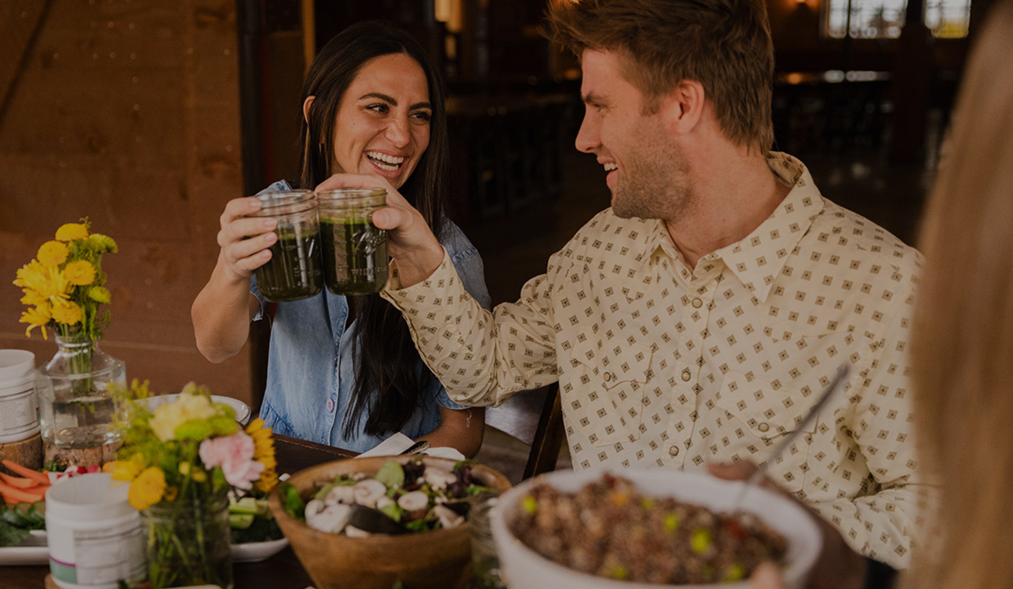 A smiling young woman and man cheers with mason jars filled with Superfueled Greens powder drinks, while sitting at a table covered in flowers and healthy salads. 