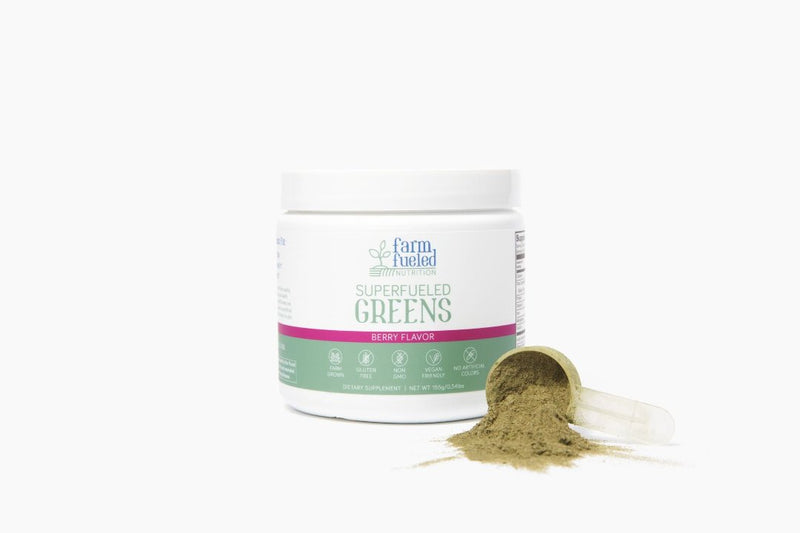 The white and green Superfueled Greens canister (Berry Flavor highlighted in pink) displayed in front of a white background, next to a clear plastic scoop overflowing with delicious greens powder in the bottom right corner.