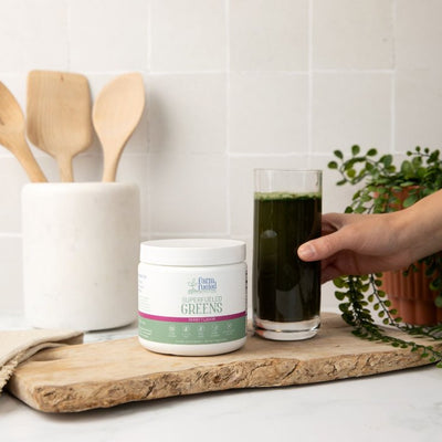 A caucasian hand grasps a tall clear glass filled with a rich dark green drink, made by mixing Superfueled Greens with water. The glass sits beside a white Superfueled Greens canister atop a rustic wooden cutting board, with cooking utensils and a plant in the background.