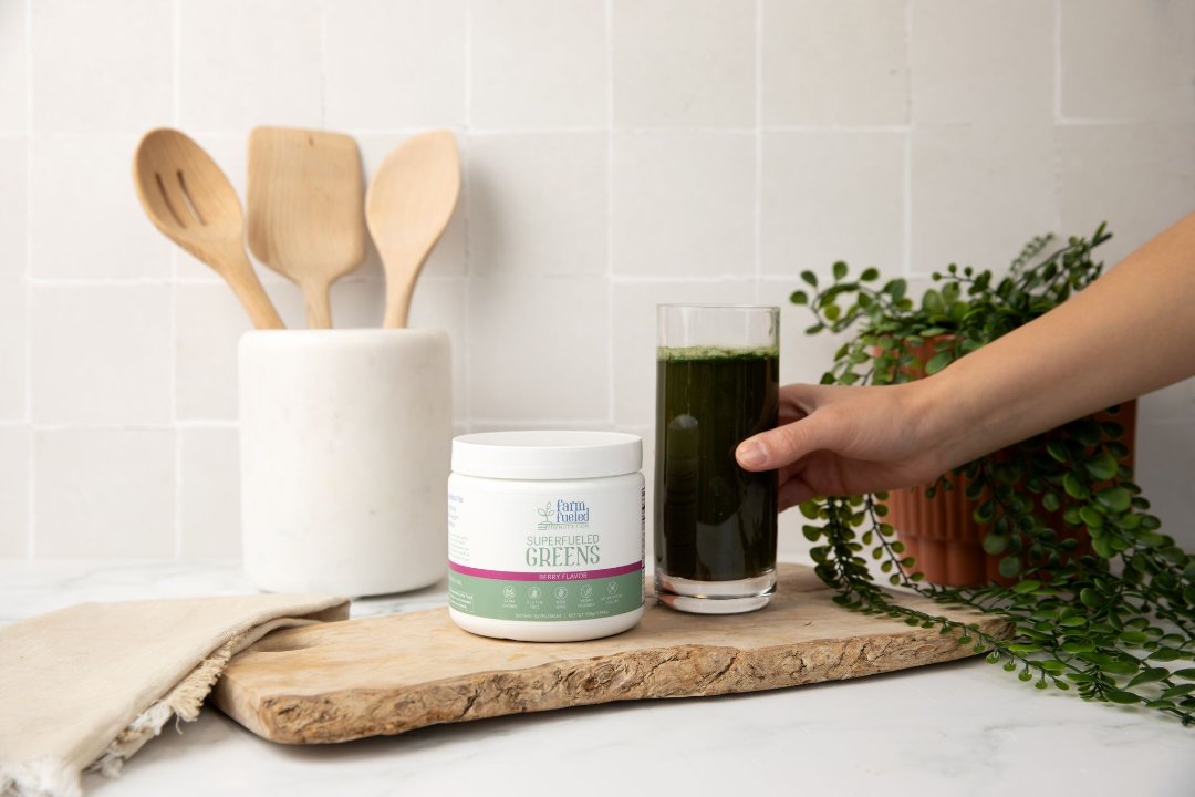 A caucasian hand grasps a tall clear glass filled with a rich dark green drink, made by mixing Superfueled Greens with water. The glass sits beside a white Superfueled Greens canister atop a rustic wooden cutting board, with cooking utensils and a plant in the background.